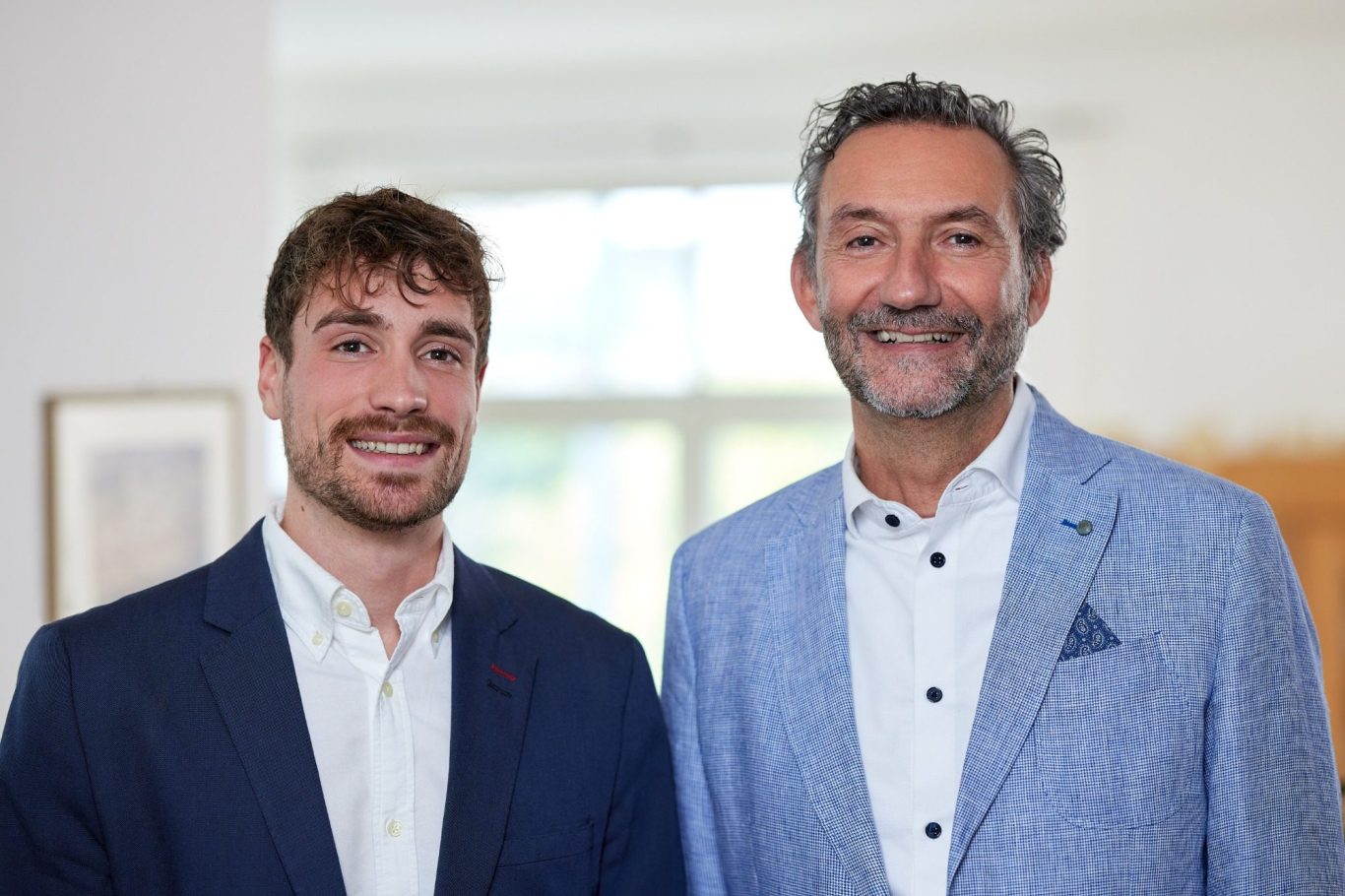 Joel und Wolfgang Frei Zwei Männer stehen lächelnd nebeneinander, einer in einem dunklen Anzug, der andere in einem hellen Blazer.